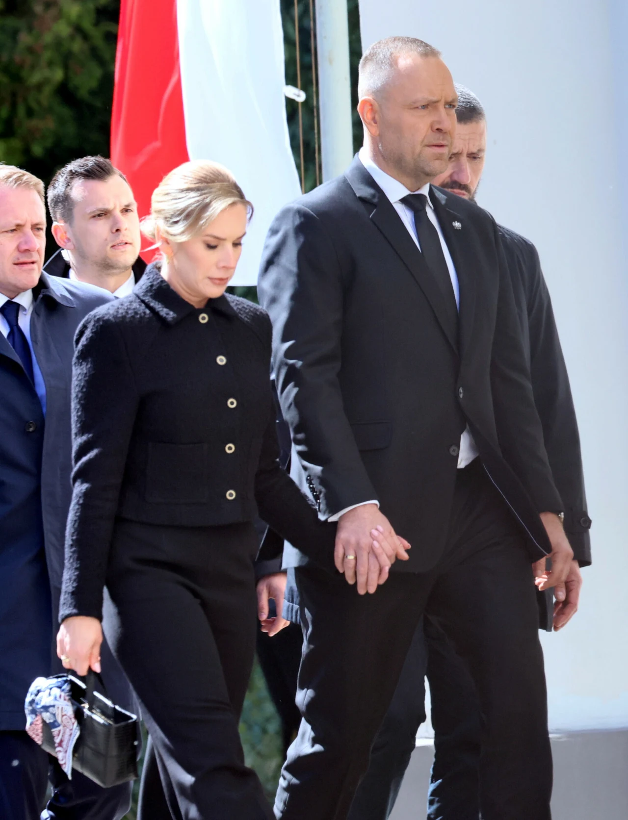 para ubrana na czarno idzie razem, trzymając się za ręce; mężczyzna ma poważny wyraz twarzy, kobieta niesie czarną torebkę, w tle widoczni są inni elegancko ubrani ludzie oraz biało-czerwony fragment flagi