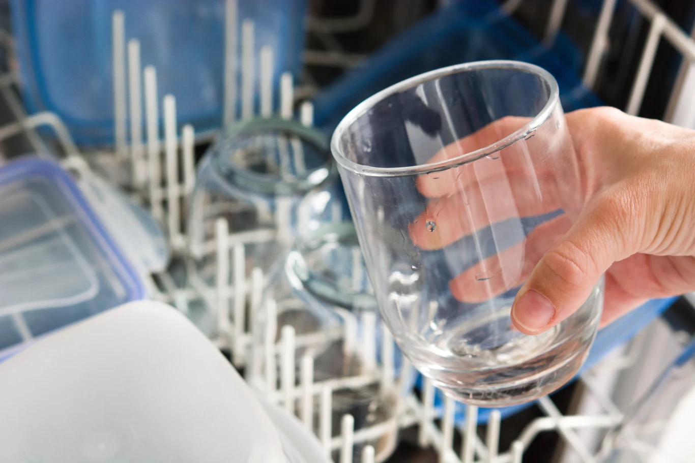 dłoń trzymająca czysty szklany kubek wyjmowany z dolnego kosza zmywarki pełnej innych naczyń i plastikowych pojemników