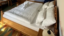 przestronny pokój z dwuosobowym łóżkiem drewnianym nakrytym białą pościelą, ręcznikiem ułożonym w kształcie łabędzia, kolorowym dywanem, drewnianą szafą, oknem z niebieskimi zasłonami, niskimi półkami na ścianie i ozdobnym malowaniem pod sufitem ze sty...