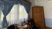 Pokój z drewnianym sufitem pomalowanym na zielono, dwoma ozdobnymi żyrandolami, dużym oknem z niebieskimi zasłonami, dwiema granatowymi fotelami przy drewnianym stoliku, na którym stoją szklanki, butelka wody i filiżanka, a w rogu stoi drewniana szafa.