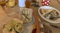 drewniany stół nakryty śniadaniem: kromki chleba z serem, szynką i ogórkiem, parówka z musztardą i keczupem, talerz z kawałkiem ciasta i łyżką, trzy szklanki z napojami oraz czerwony kubek w białe kropki; w tle stylowa kuchnia z półkami pełnymi porcela...