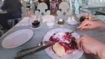 bogato zdobiona sala restauracyjna z jasnoniebieskimi drewnianymi elementami i malowanymi sufitami, w centrum stołu talerz z deserem polanym sosem owocowym, dłoń trzymająca widelec, obok filiżanki, szklanki i przyprawy, w tle osoby siedzące przy stołach