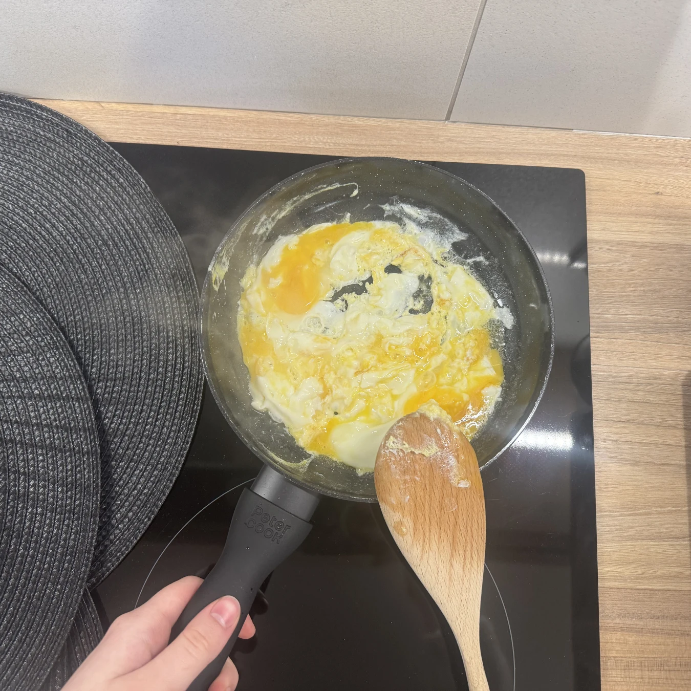 Moja tania patelnia z Biedronki patelnia z częściowo ściętymi jajkami mieszanymi drewnianą łyżką nad płytą indukcyjną, obok widoczne okrągłe podkładki