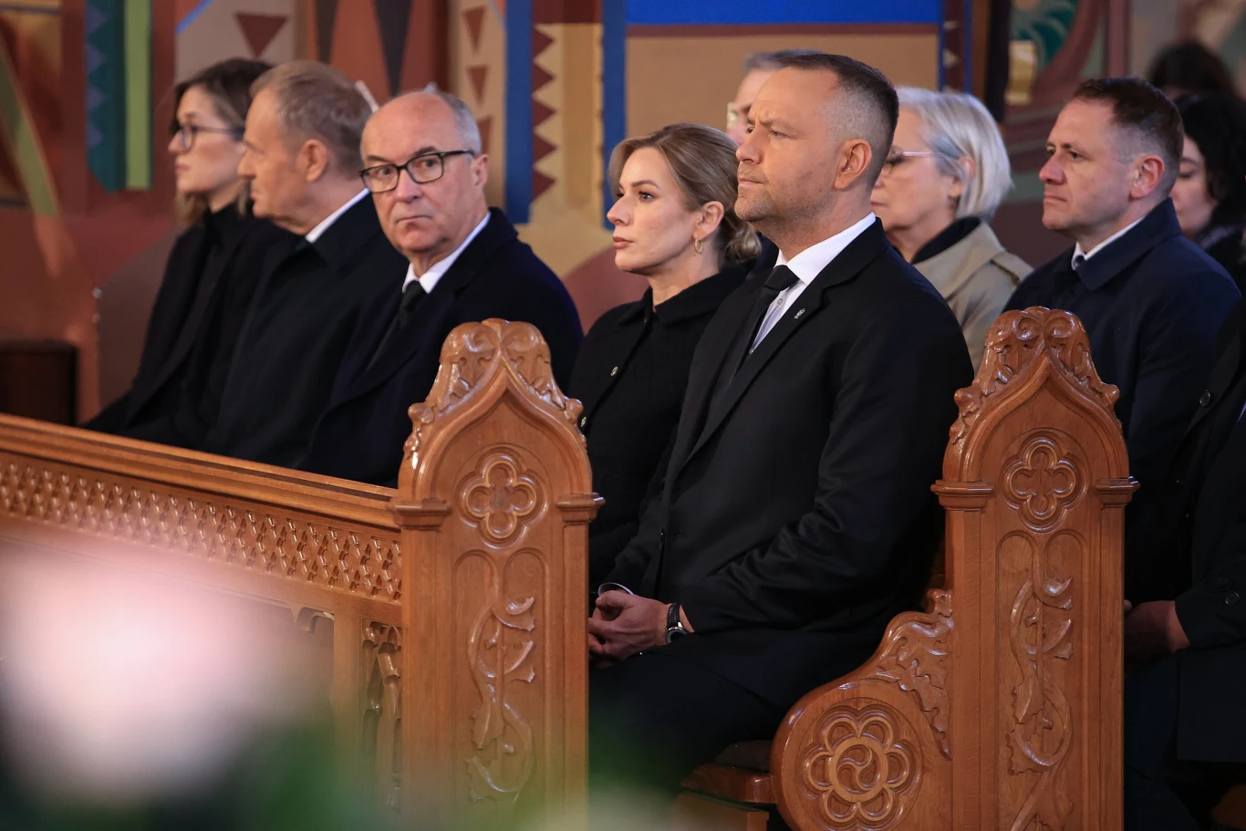 Najważniejsze osoby w państwie na pogrzebie Łukasza Litewki Grupa elegancko ubranych osób siedzi w pierwszych ławkach kościoła, skupieni i zamyśleni, otoczeni dekoracyjnymi, drewnianymi elementami wnętrza świątyni.
