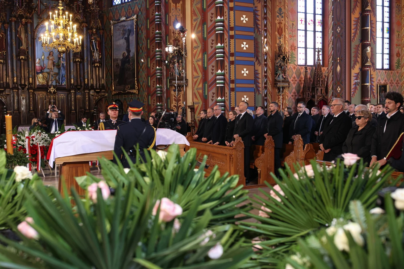 Pogrzeb Łukasza Litewki - wnętrze kościoła trumna nakryta białym materiałem z czerwonym paskiem ustawiona w ozdobnym kościele, wokół niej zgromadzeni żałobnicy ubrane na czarno, w tle bogato zdobiony ołtarz i kolorowe witraże, z przodu dekoracje z zielonych liści i różowych kwiatów