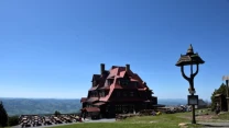 drewniane schronisko górskie o charakterystycznej czerwonej mansardowej konstrukcji, stojące na otwartej polanie z ławkami dla turystów, w tle rozległe górskie panoramy pod błękitnym niebem oraz drewniana kapliczka po prawej stronie
