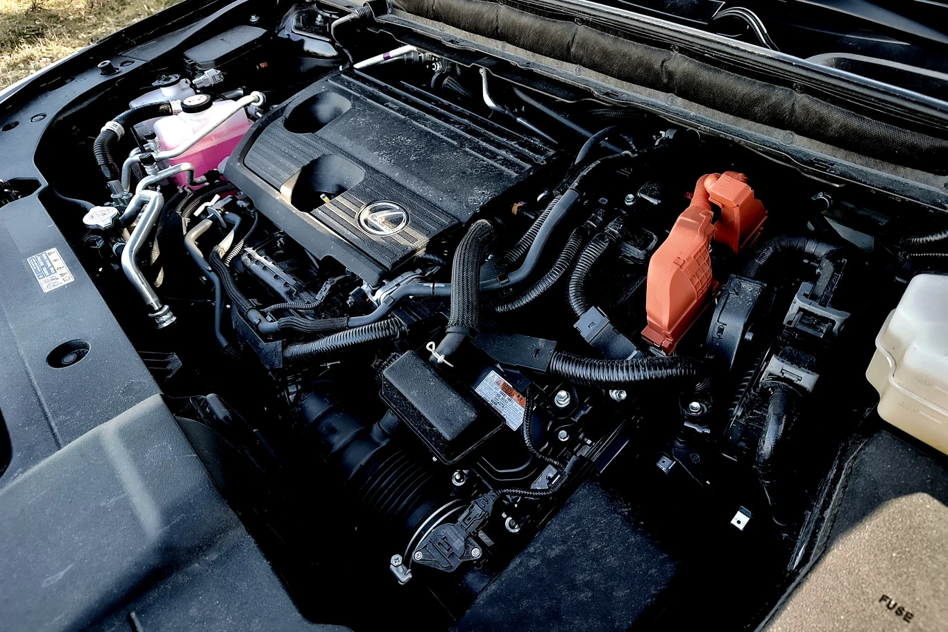 silnik hybrydowy marki Lexus z wyeksponowanymi przewodami i osłonami, charakterystyczną pomarańczową obudową podzespołu elektrycznego oraz logo producenta widocznym na pokrywie silnika