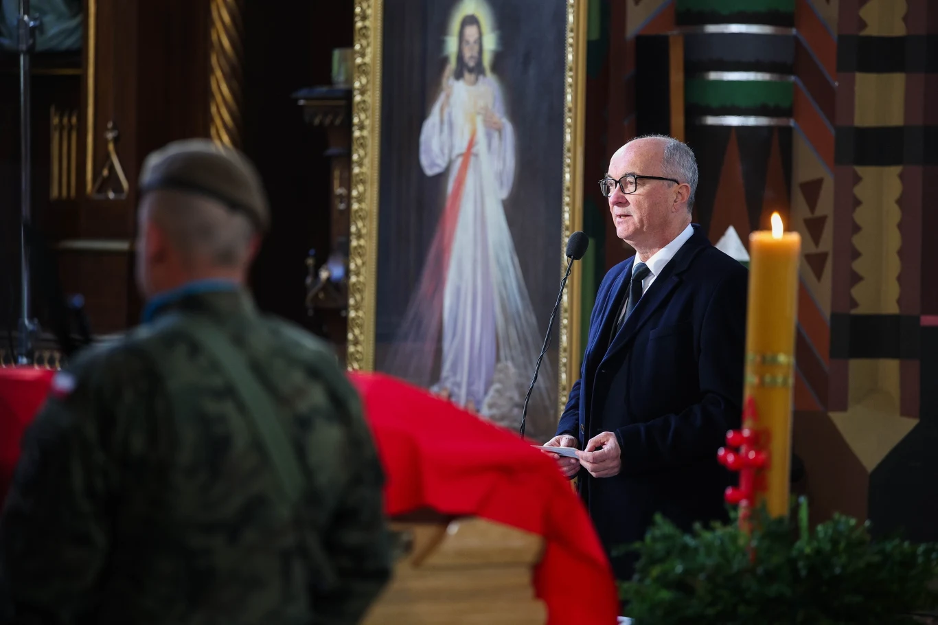 Marszałek Sejmu Włodzimierz Czarzasty na pogrzebie Łukasza Litewki mężczyzna w garniturze wygłasza przemówienie przy mikrofonie w kościele, obok znajduje się trumna przykryta czerwoną flagą, z przodu żołnierz w mundurze, w tle duży obraz Jezusa Miłosiernego i zapalona paschałka z czerwoną dekoracją