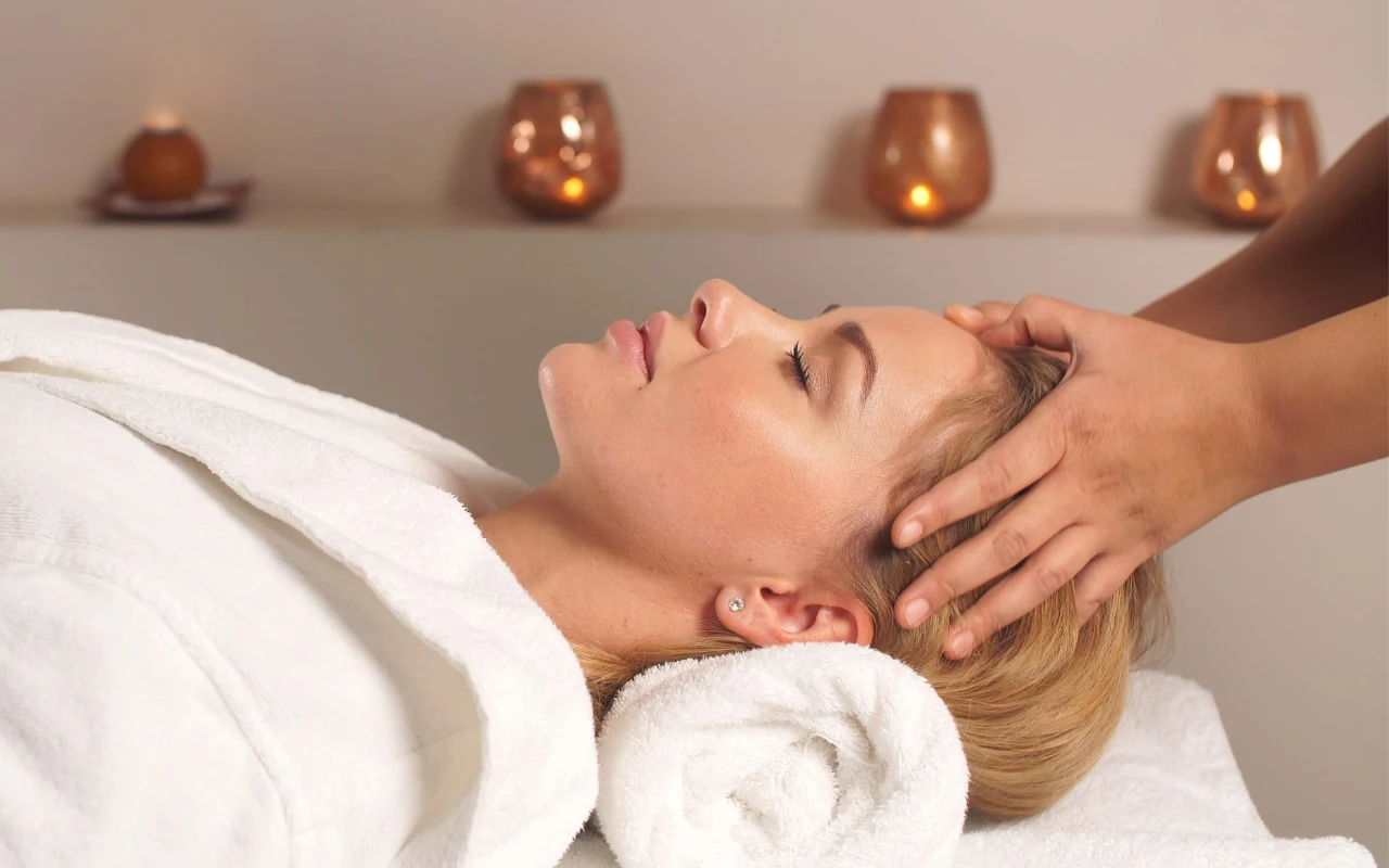 kobieta w białym szlafroku leży na plecach z głową opartą na białym ręczniku, a druga osoba wykonuje jej relaksacyjny masaż głowy; w tle płoną świece tworzące przytulną atmosferę