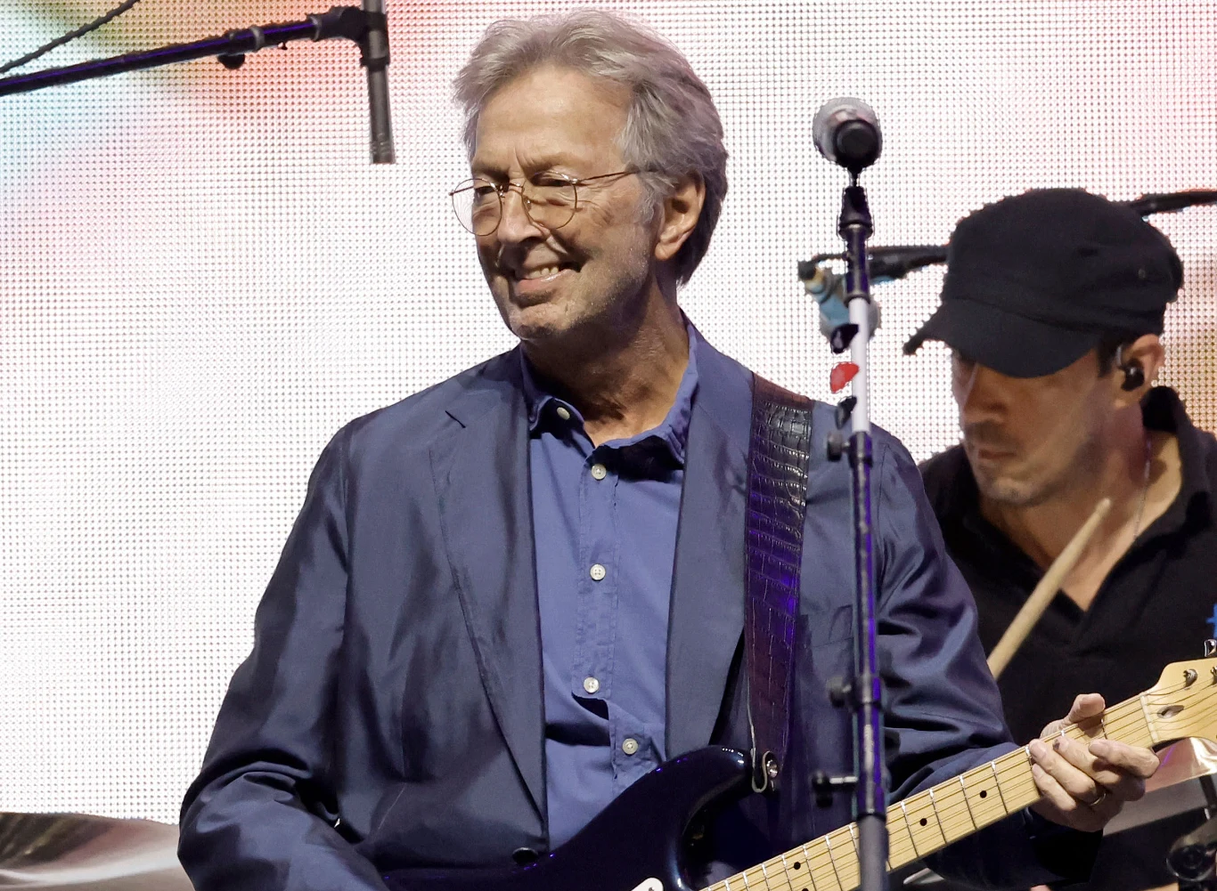 Eric Clapton nazywany jest "bogiem gitary" Eric Clapton w okularach i niebieskiej marynarce gra na gitarze elektrycznej; za nim perkusista na oświetlonej scenie.