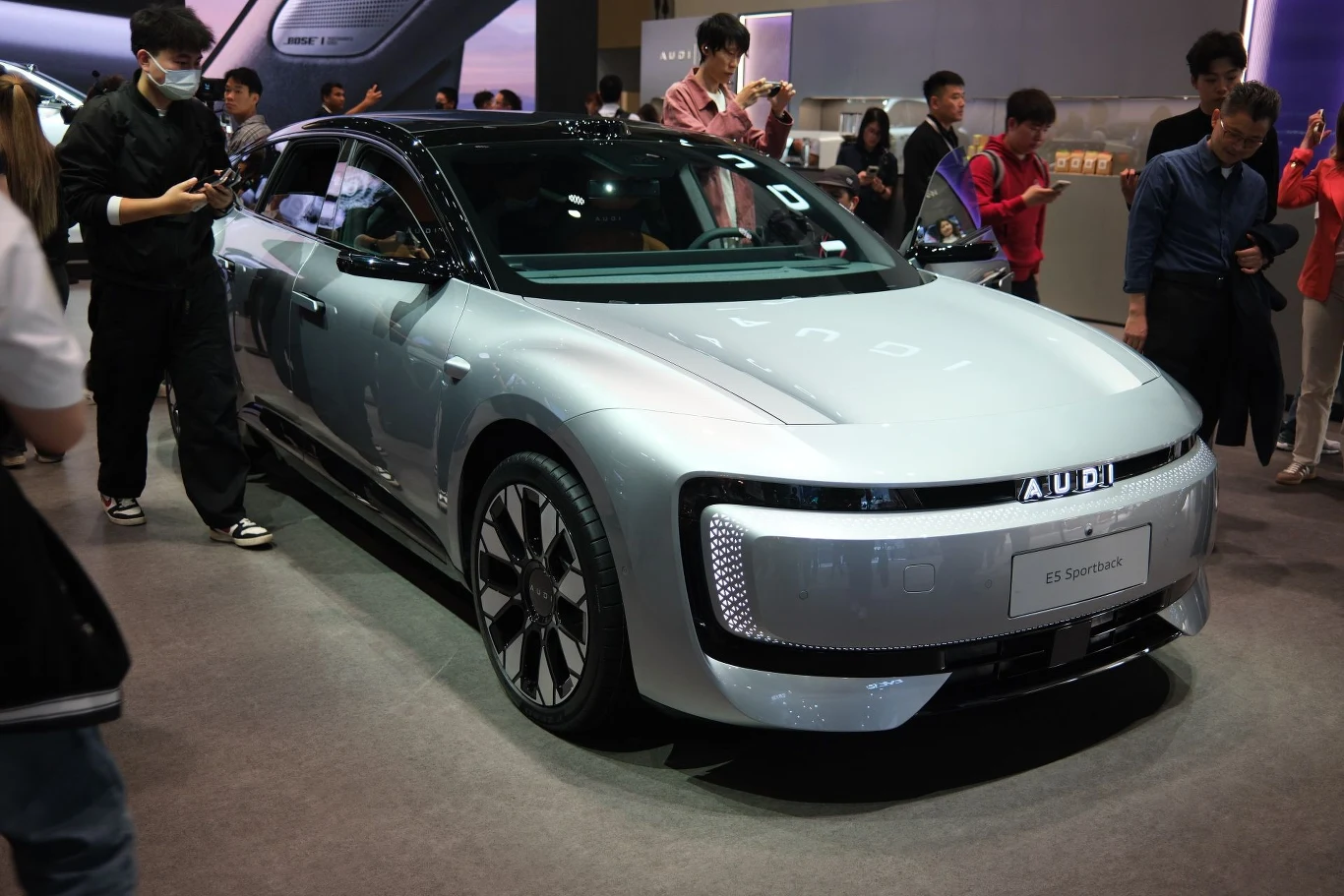 Za stylistykę AUDI E5 Sportback odpowiada Kamil Łabanowicz. nowoczesny, srebrny samochód elektryczny Audi z futurystycznymi reflektorami LED i czarnym dachem stoi na wystawie, otoczony przez ludzi fotografujących i obserwujących pojazd