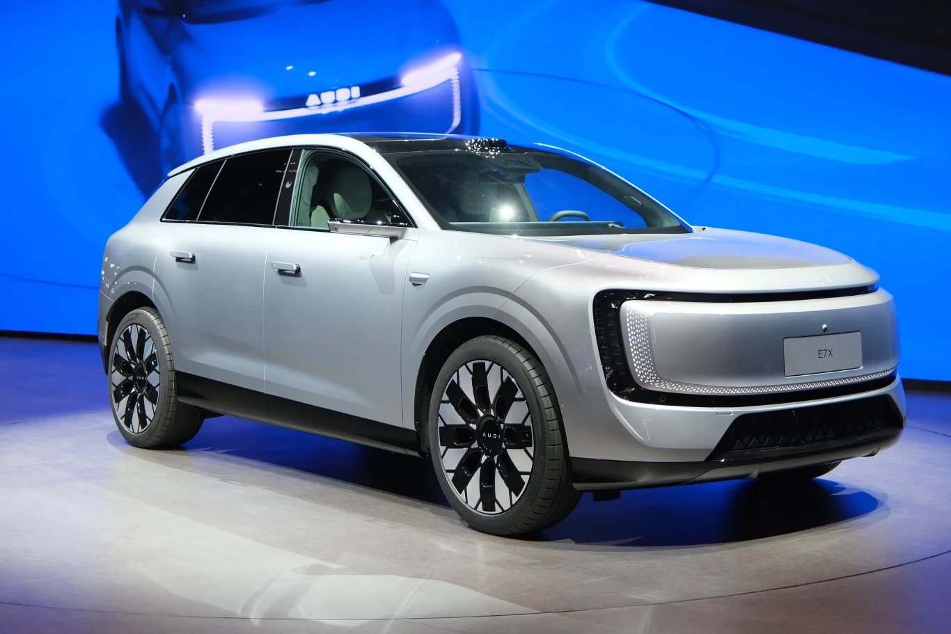 E7X to pierwszy SUV submarki AUDI. srebrny samochód elektryczny typu SUV o nowoczesnym designie, dużych felgach i minimalistycznym przodzie z podświetlanymi elementami, ustawiony na podświetlonej platformie wystawowej przed ekranem prezentującym jego sylwetkę