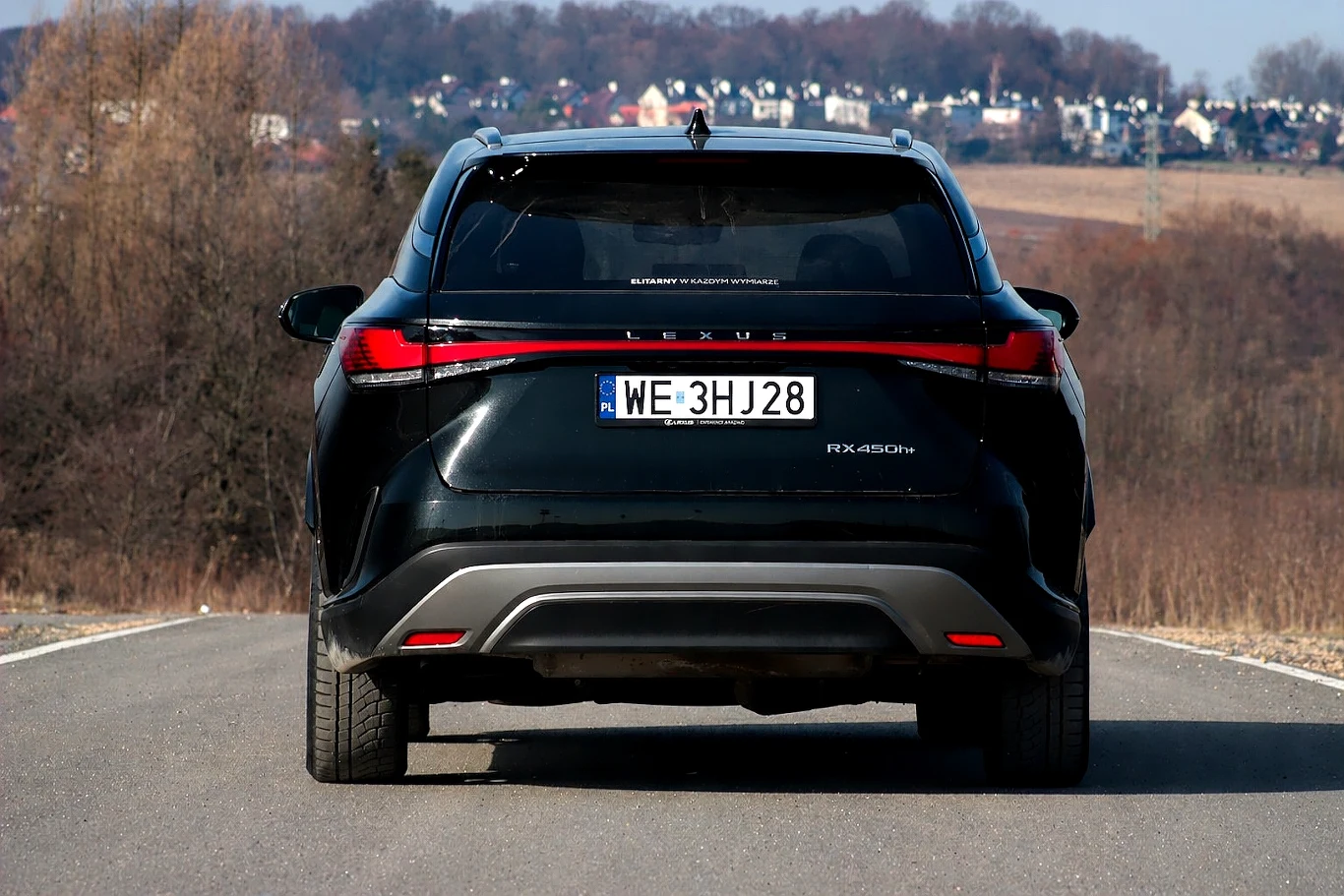 czarny samochód typu SUV marki Lexus RX 450h, widziany od tyłu, stoi na asfaltowej drodze na tle drzew oraz oddalonego osiedla domów