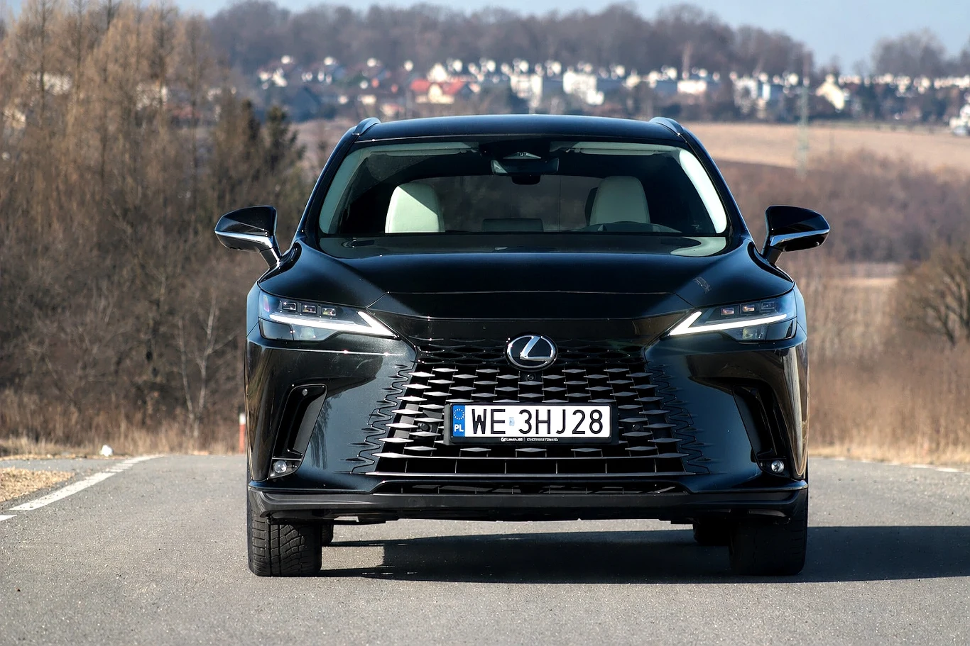 Czarny samochód marki Lexus z wyraźnie widocznym dużym przednim grillem, nowoczesnymi reflektorami oraz polską tablicą rejestracyjną, ustawiony centralnie na asfaltowej drodze, w tle rozległy krajobraz z polami i zabudowaniami na horyzoncie.