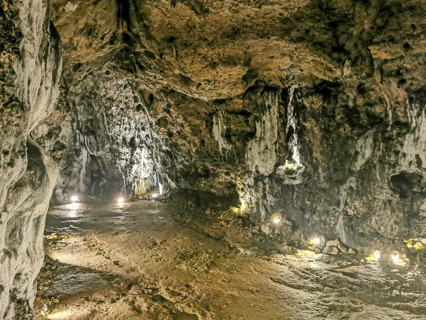 Skalna grota o nieregularnych ścianach i sklepieniu, rozświetlona subtelnym światłem umieszczonym przy ziemi, z widocznymi naciekami mineralnymi i wilgotnym, nierównym podłożem.