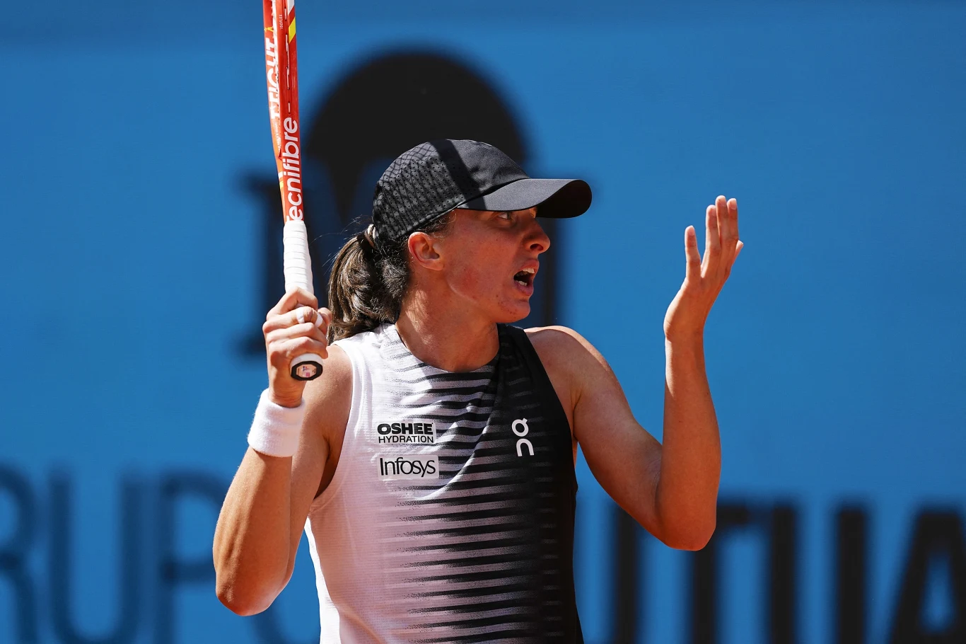 tenisistka w sportowym stroju i czapce z daszkiem trzyma w dłoni rakietę tenisową, gestykulując drugą ręką z wyrazem frustracji na twarzy, na tle niebieskiego kortu