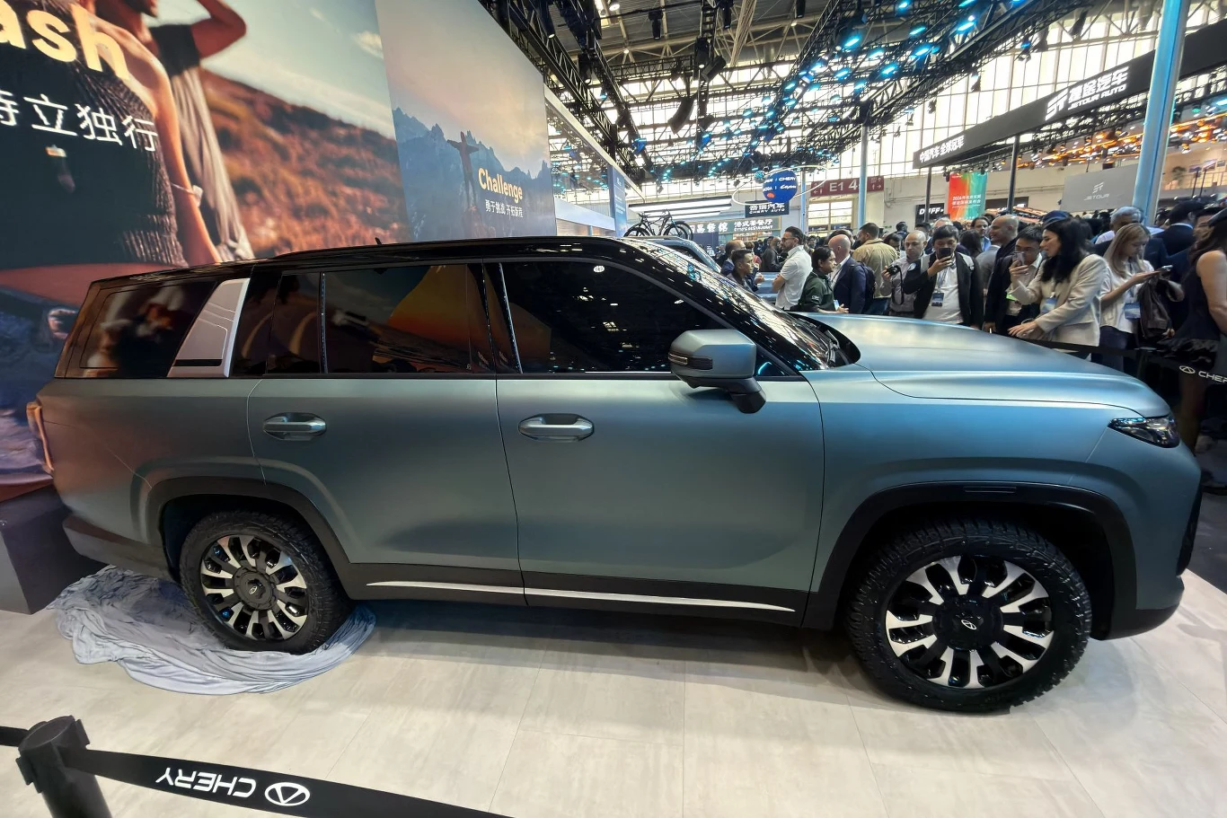 Chery Tiggo V może być siedmioosobowym SUV-em, ale w razie potrzeby można zdemontować siedzenia trzeciego rzędu i dach od słupka C. Wówczas auto staje się pick-upem. duży, nowoczesny SUV w kolorze matowej szarości prezentowany na targach motoryzacyjnych, na tle tłumu odwiedzających i jasnego, przestronnego wnętrza hali