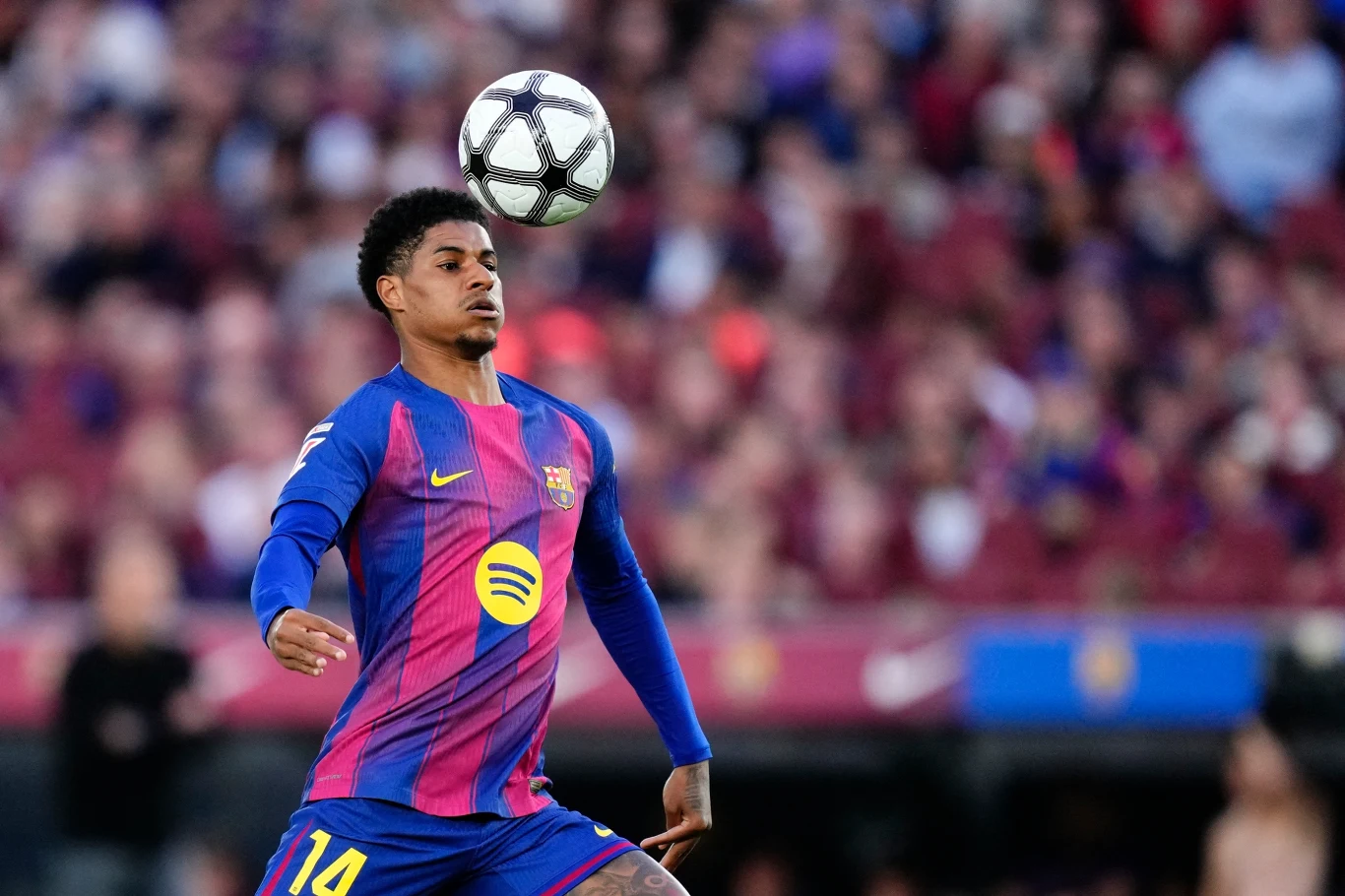 piłkarz w koszulce FC Barcelony z numerem 14 skupiony kontroluje piłkę głową podczas meczu, na tle stadionu wypełnionego kibicami