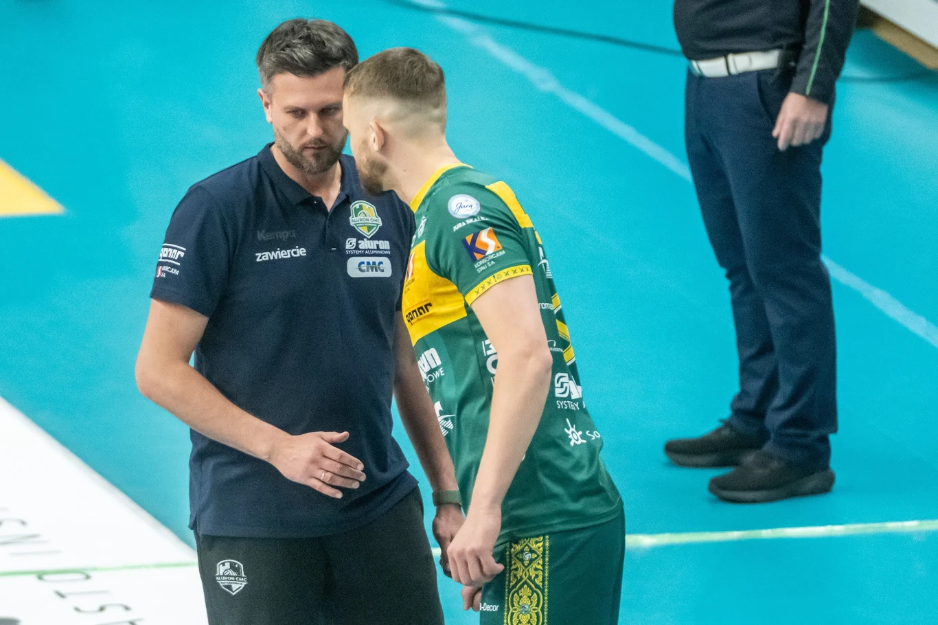 trener drużyny siatkówki w granatowej koszulce rozmawia z zawodnikiem w zielono-żółtym stroju, obaj skoncentrowani i zaangażowani, stoją blisko siebie na boisku sportowym