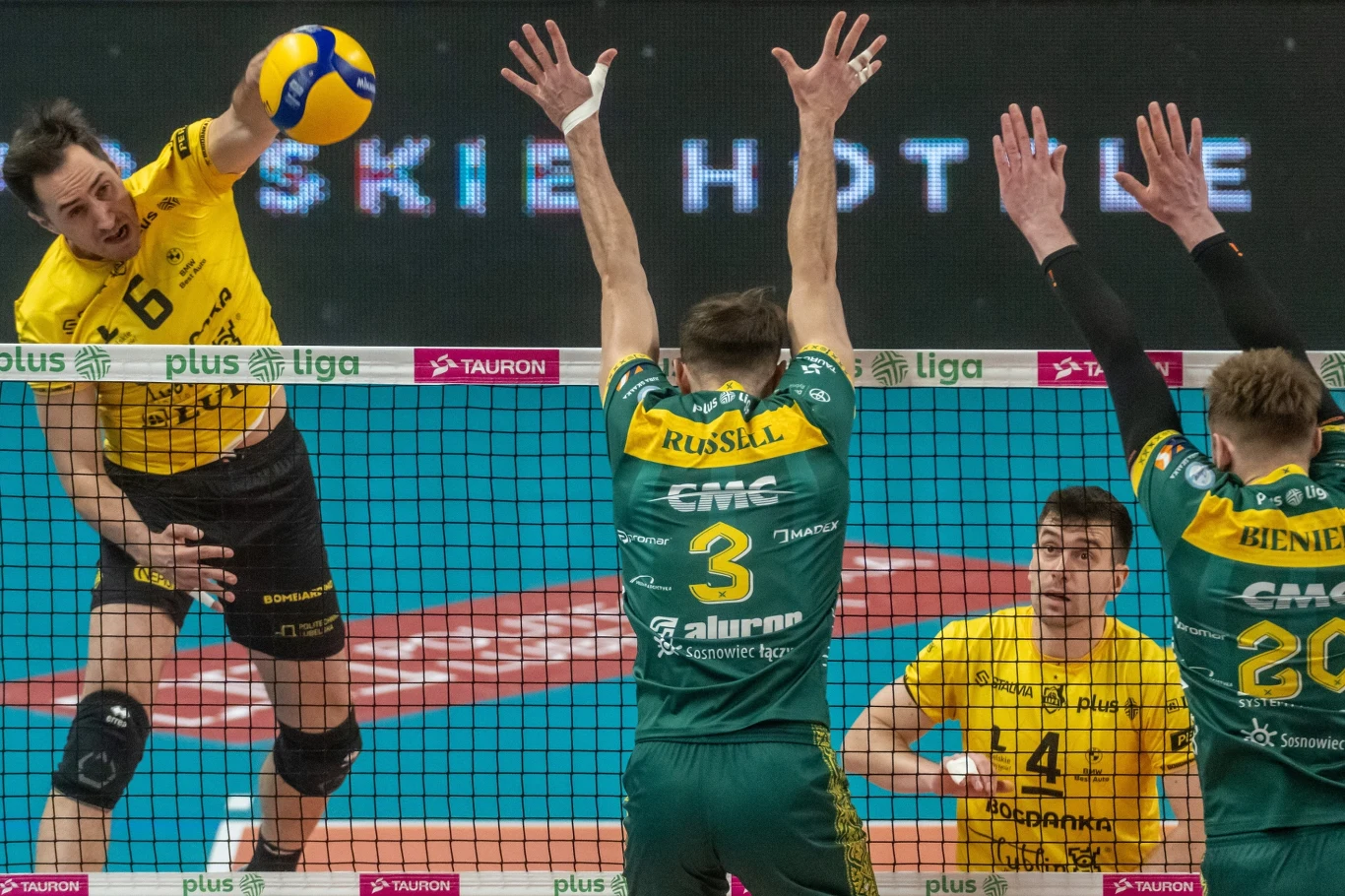 W ataku Mateusz Malinowski z Bogdanki LUK Lublin zawodnik w żółtej koszulce wykonuje atak z lewego skrzydła, piłka znajduje się tuż nad siatką, dwaj siatkarze w zielonych koszulkach próbują zablokować atak, a po prawej stronie widoczny jest rozgrywający w żółtej koszulce obserwujący akcję