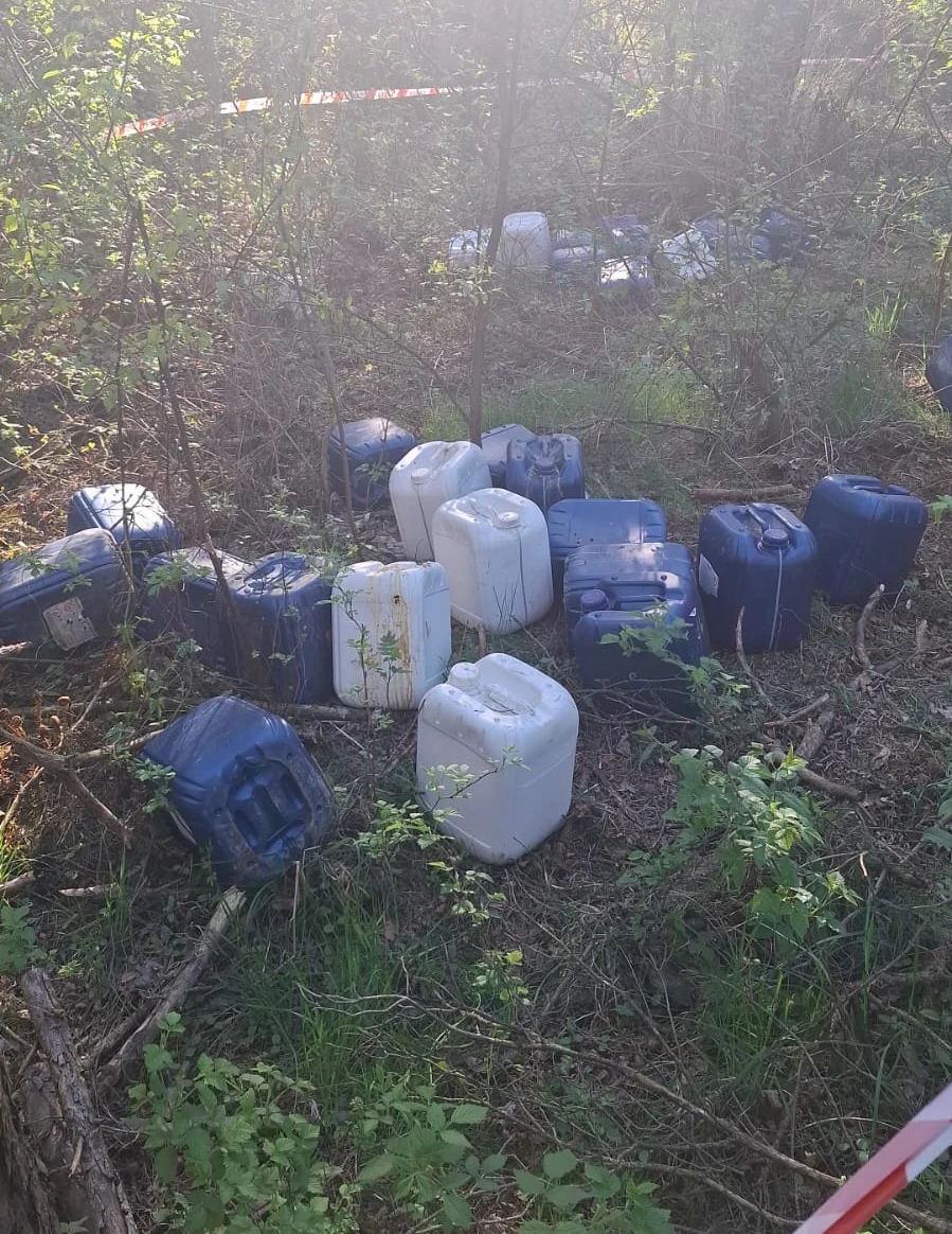 Porozrzucane w zaroślach niebieskie i białe kanistry plastikowe otoczone gęstą roślinnością, gdzieniegdzie widoczna taśma ostrzegawcza.