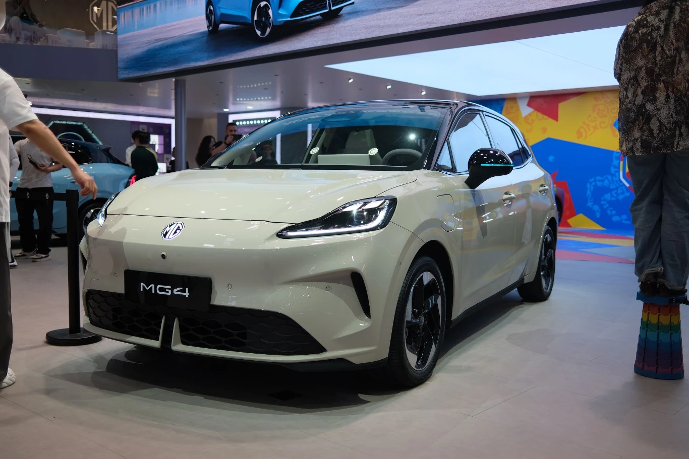 Nowe MG4, w Europie z dopiskiem "Urban", różni się od dotychczasowego MG4 np. tym, że ma napęd na przednią, a nie na tylną oś. biały samochód elektryczny MG4 ustawiony na wystawie motoryzacyjnej, zwrócony przodem z widocznymi reflektorami LED, czarnymi felgami i nowoczesnym, aerodynamicznym kształtem nadwozia