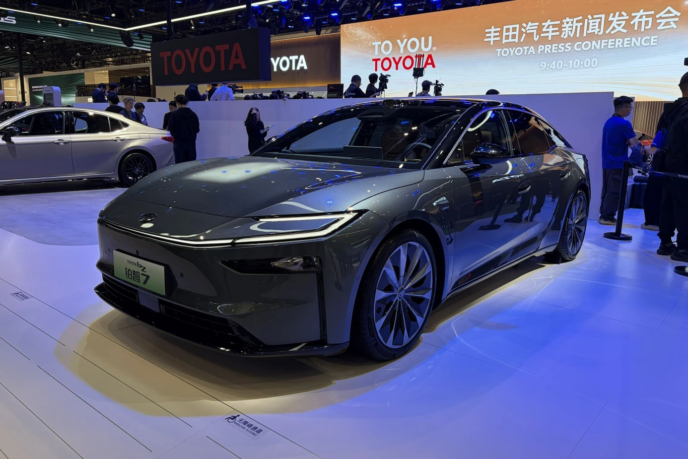 Toyota bZ7 jest większa od Tesli Model S, ale tańsza od Modelu 3. nowoczesny, szary samochód elektryczny marki Nio stoi na wystawie motoryzacyjnej oświetlony reflektorami, w tle widoczni są ludzie oraz duże logo Toyota i informacje o konferencji prasowej na jasnym ekranie