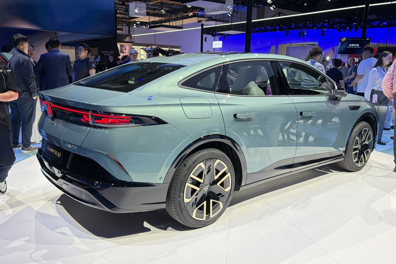 Volkswagen ID.Unyx 07 został zbudowany na platformie opracowanej wspólnie z firmą XPeng. Nowoczesny, zielony crossover coupe o sportowej sylwetce z wyrazistymi tylnymi światłami LED i dwukolorowymi felgami, prezentowany w jasnym salonie wystawowym, w otoczeniu zwiedzających.