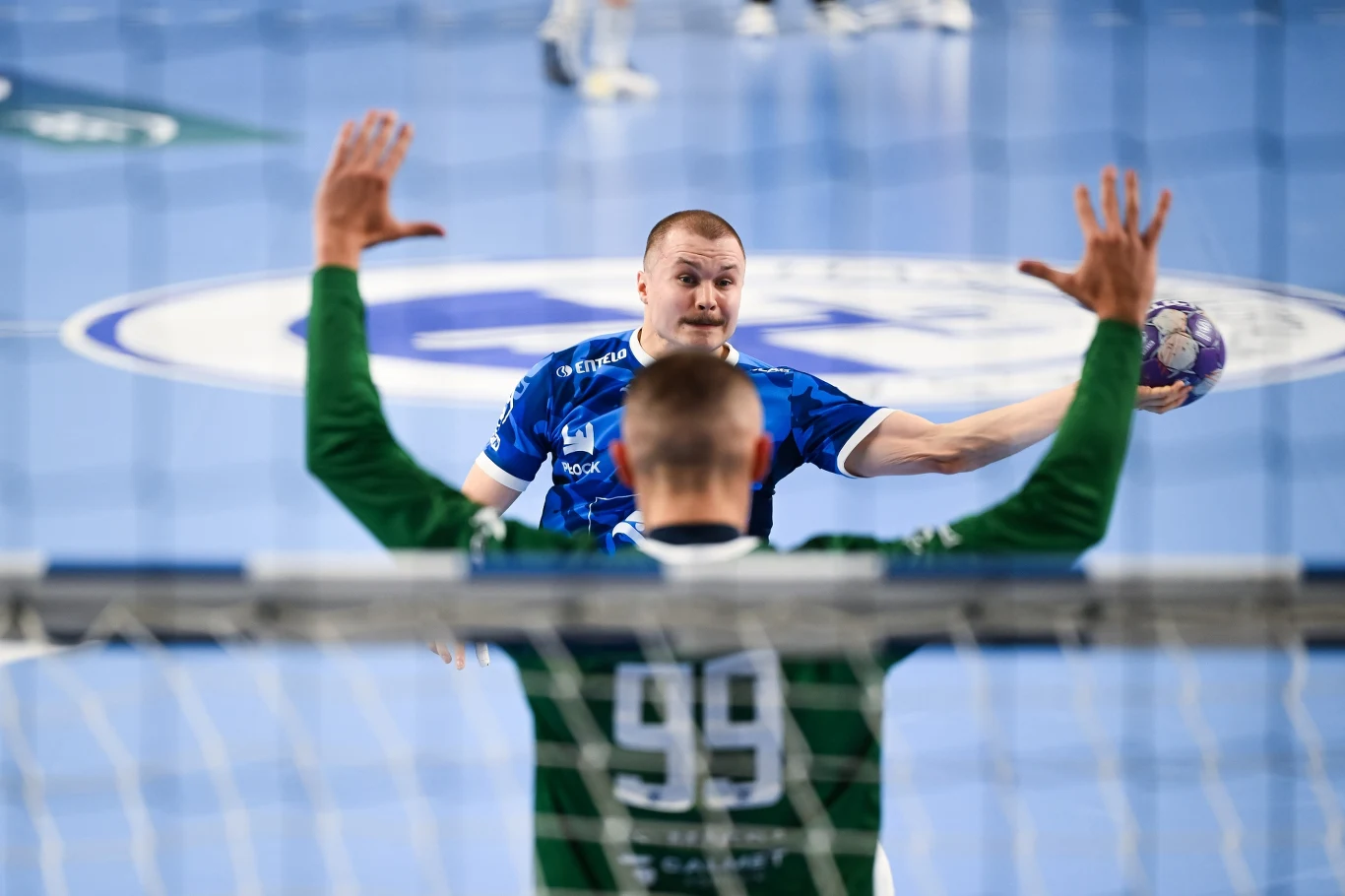 zawodnik w niebieskiej koszulce przygotowujący się do rzutu piłką ręczną w kierunku bramkarza w zielonym stroju z numerem 99, obaj skupieni na akcji tuż przed bramką