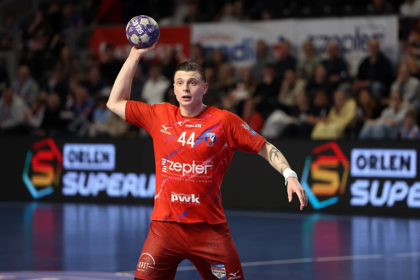 zawodnik drużyny piłki ręcznej w czerwonym stroju z numerem 44 trzyma piłkę nad głową, gotowy do rzutu, na tle trybun wypełnionych kibicami oraz widocznych band reklamowych Orlen Superliga