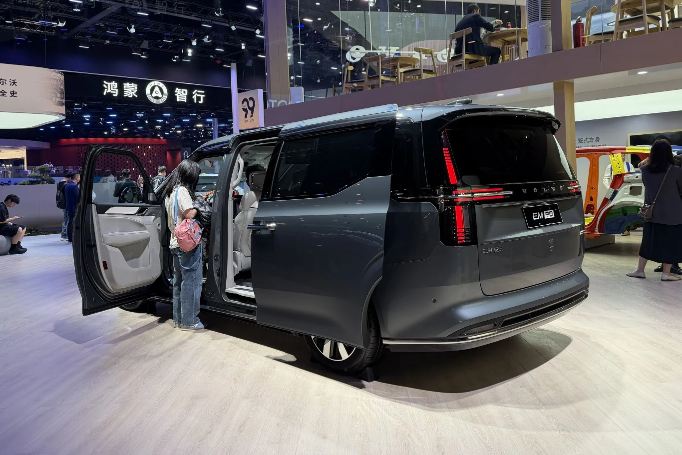 Volvo EM90 to elektryczny luksusowy van. ciemnoszary minivan z otwartymi drzwiami bocznymi, przy którym stoi osoba z różowym plecakiem zaglądająca do środka, nowoczesne oświetlenie wnętrza targów samochodowych w tle