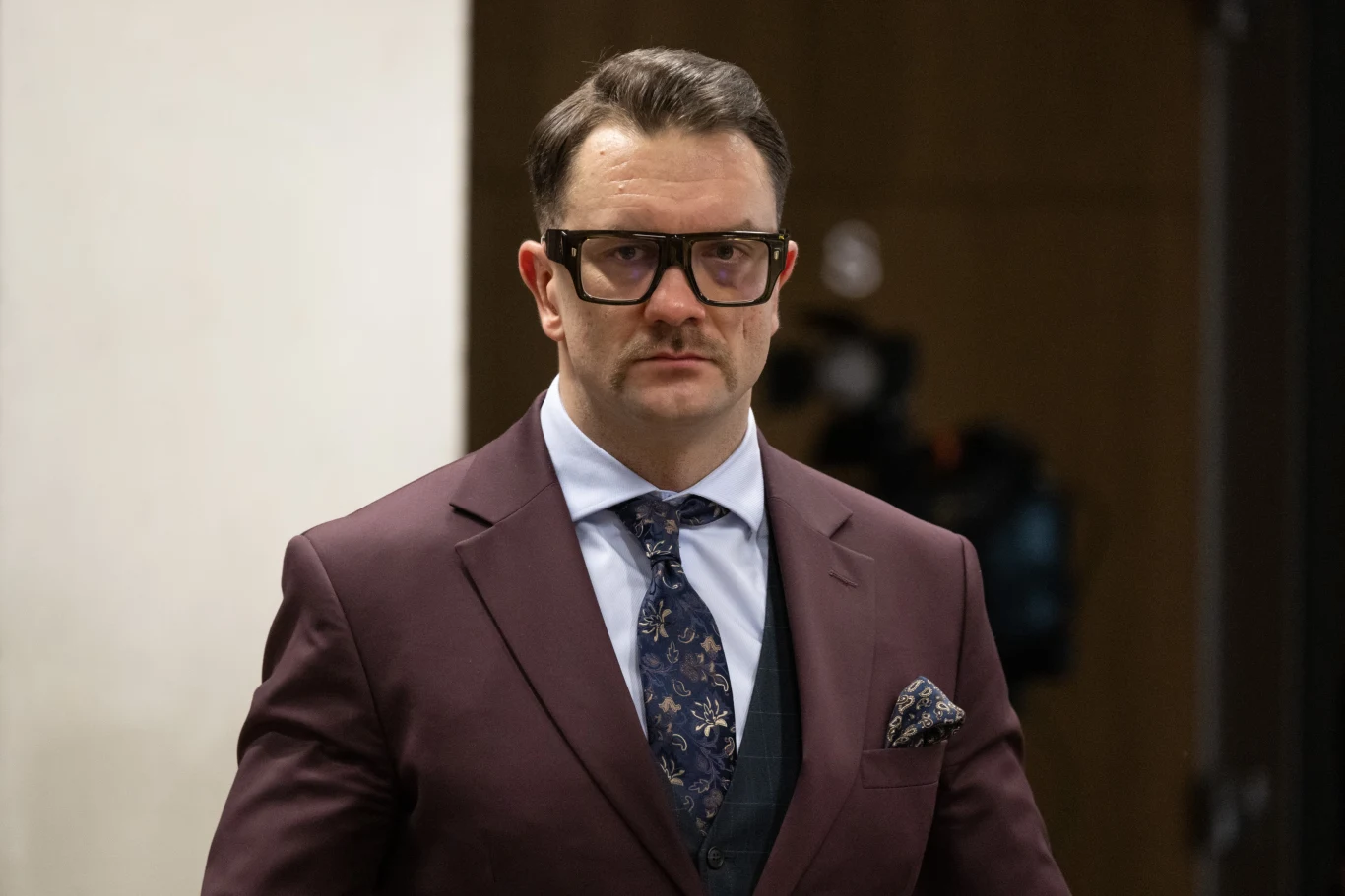 Łukasz Mejza Łukasz Mejza w bordowym garniturze, jasnej koszuli i okularach, stoi w formalnym wnętrzu przy sprzęcie technicznym.