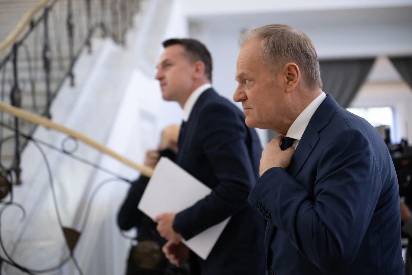 Święto Pracy dniem rozliczeń. Czy rząd Donalda Tuska ma sobie coś do zarzucenia? Donald Tusk i Adam Szłapka w garniturach na tle schodów; jeden poprawia krawat, drugi trzyma dokumenty.