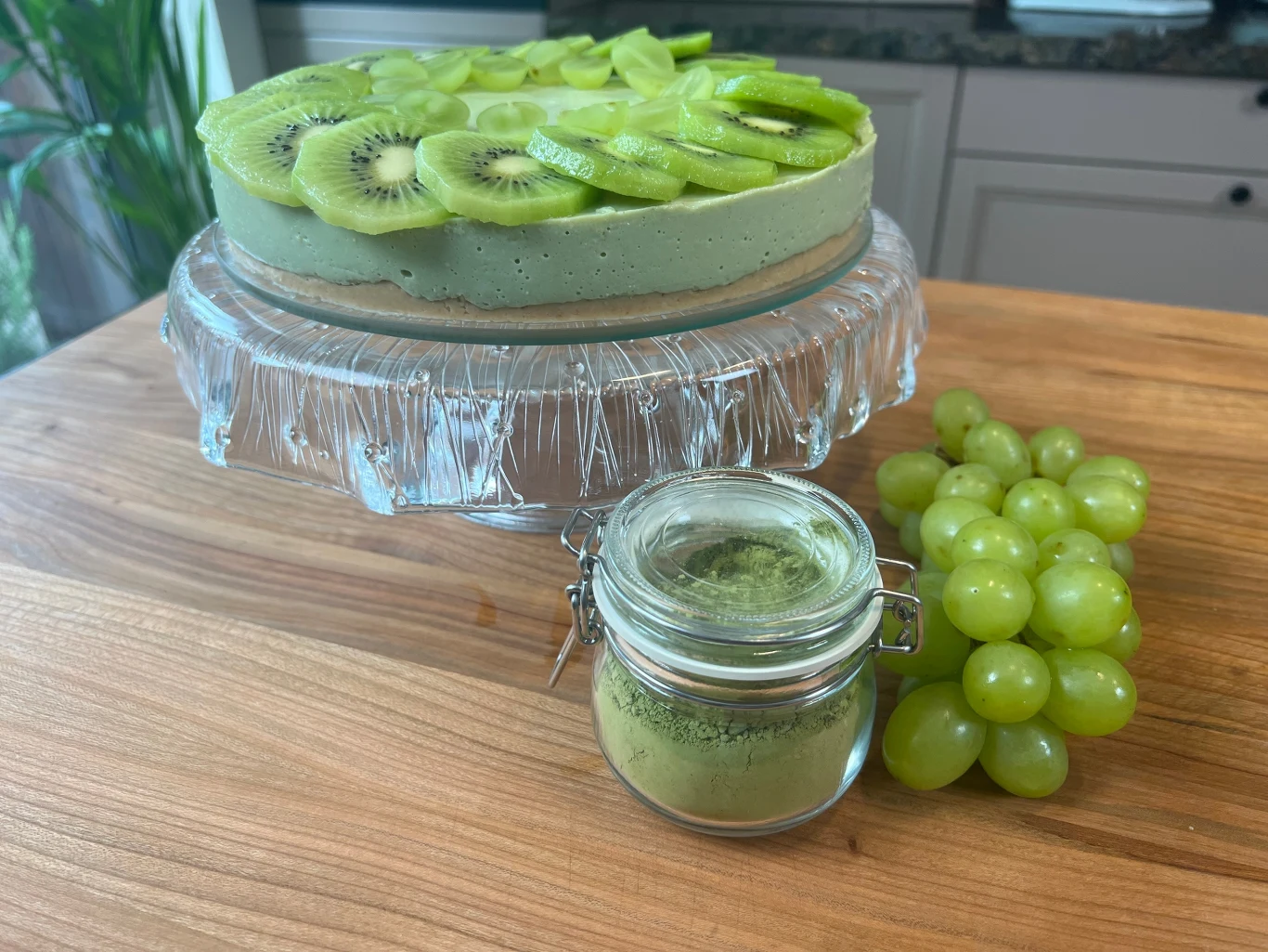 Torcik z zielonym kremem udekorowany plasterkami kiwi, ustawiony na szklanej paterze, obok kiść zielonych winogron oraz słoiczek z zielonym proszkiem matcha na drewnianym blacie.