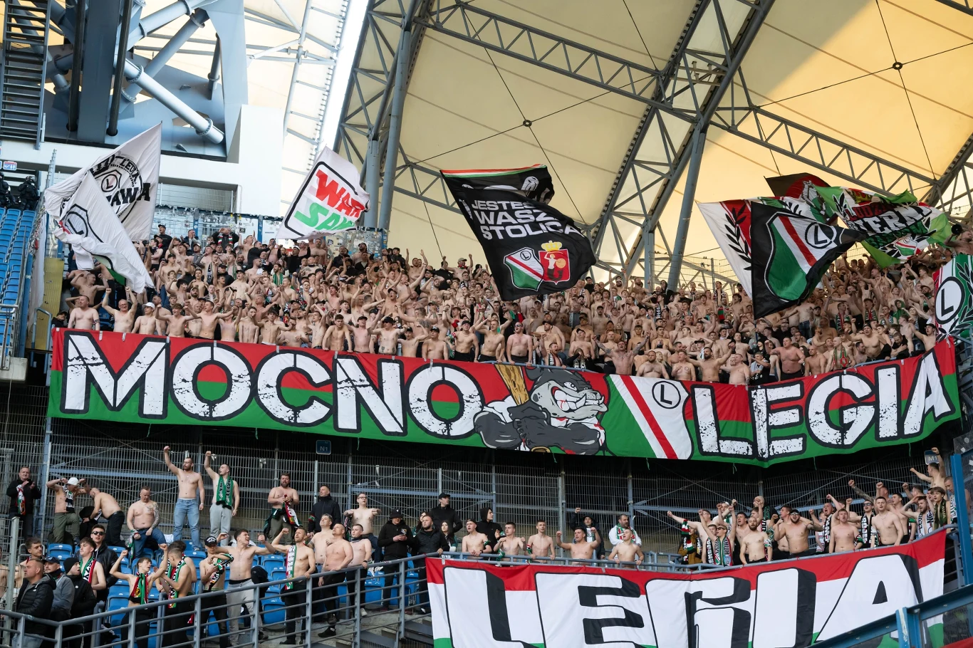 liczna grupa kibiców Legii Warszawa na stadionie, machających flagami i transparentami w barwach klubu, ubrani głównie w klubowe gadżety lub bez koszulek, wyrażający swoje wsparcie i emocje poprzez gesty oraz śpiew