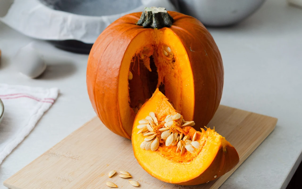 Pomarańczowa dynia z wyciętą ćwiartką leżąca na desce do krojenia, widoczne pestki zarówno w wyciętym kawałku, jak i w środku warzywa.