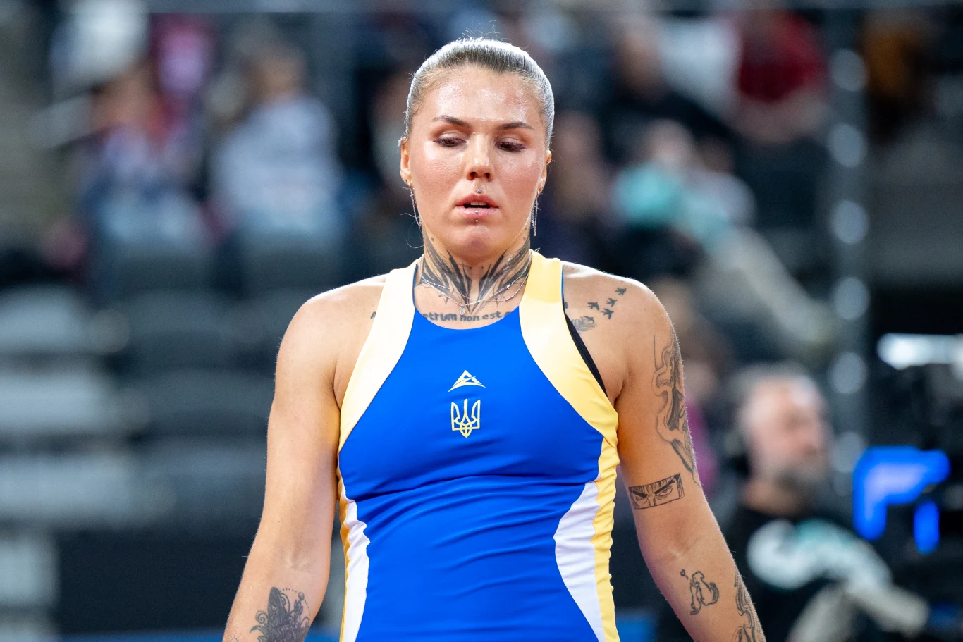 Ołeksandra Olijnykowa zawodniczka w stroju sportowym w barwach Ukrainy z widocznymi tatuażami na ramionach i szyi, skupiona na arenie sportowej