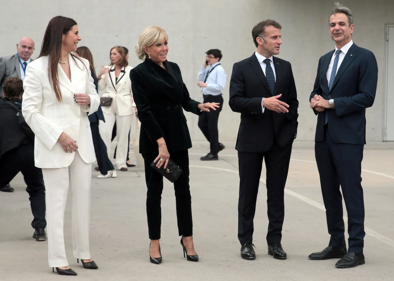 Cztery elegancko ubrane osoby stojące na otwartej przestrzeni, dwie kobiety w jasnym i ciemnym garniturze oraz dwóch mężczyzn w granatowych garniturach prowadzących rozmowę; pozostali ludzie w tle występują w formalnych strojach.