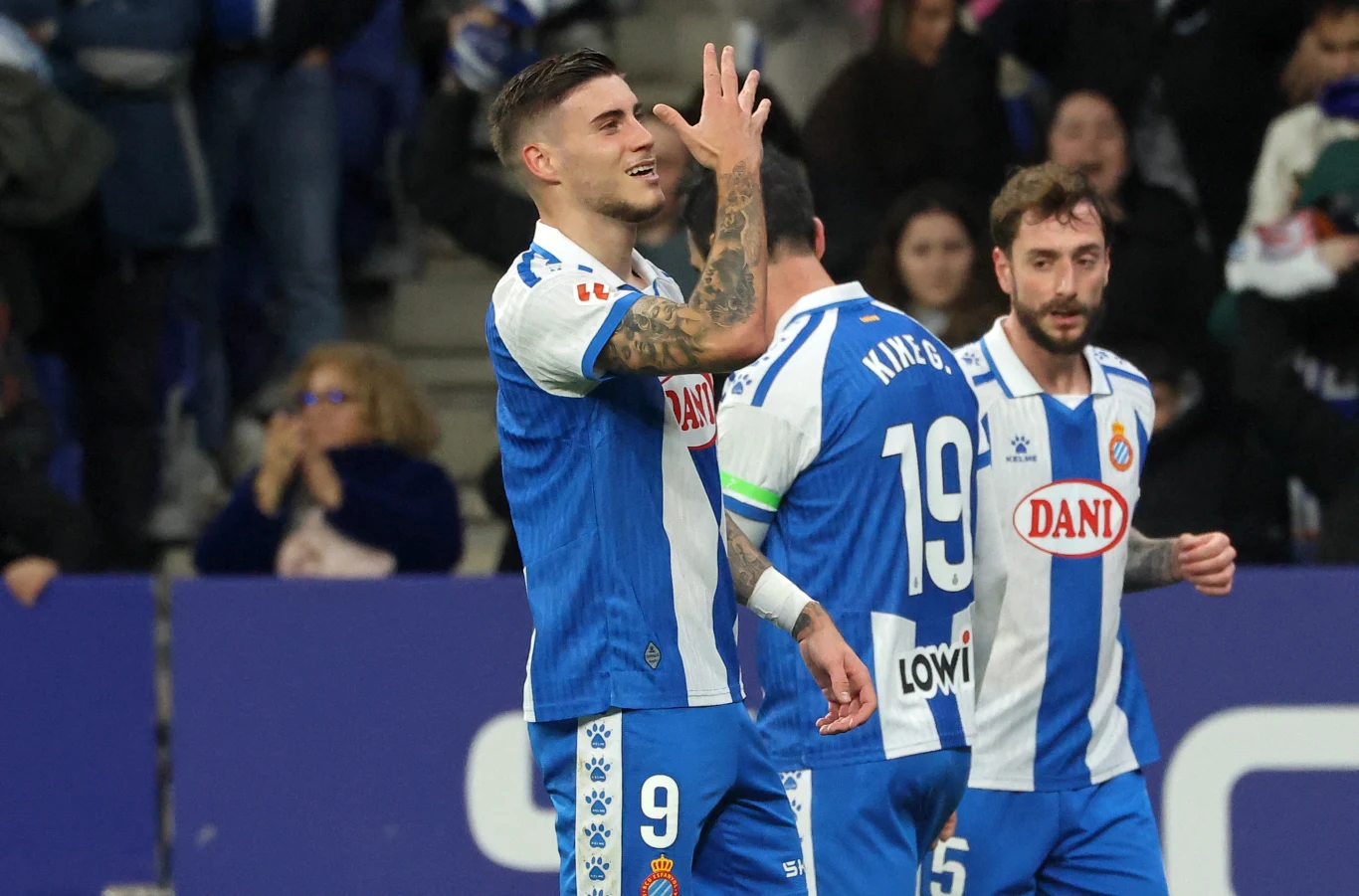trzech piłkarzy w biało-niebieskich strojach drużyny Espanyol świętuje zdobycie gola na murawie stadionu, wokół nich widać kibiców na trybunach
