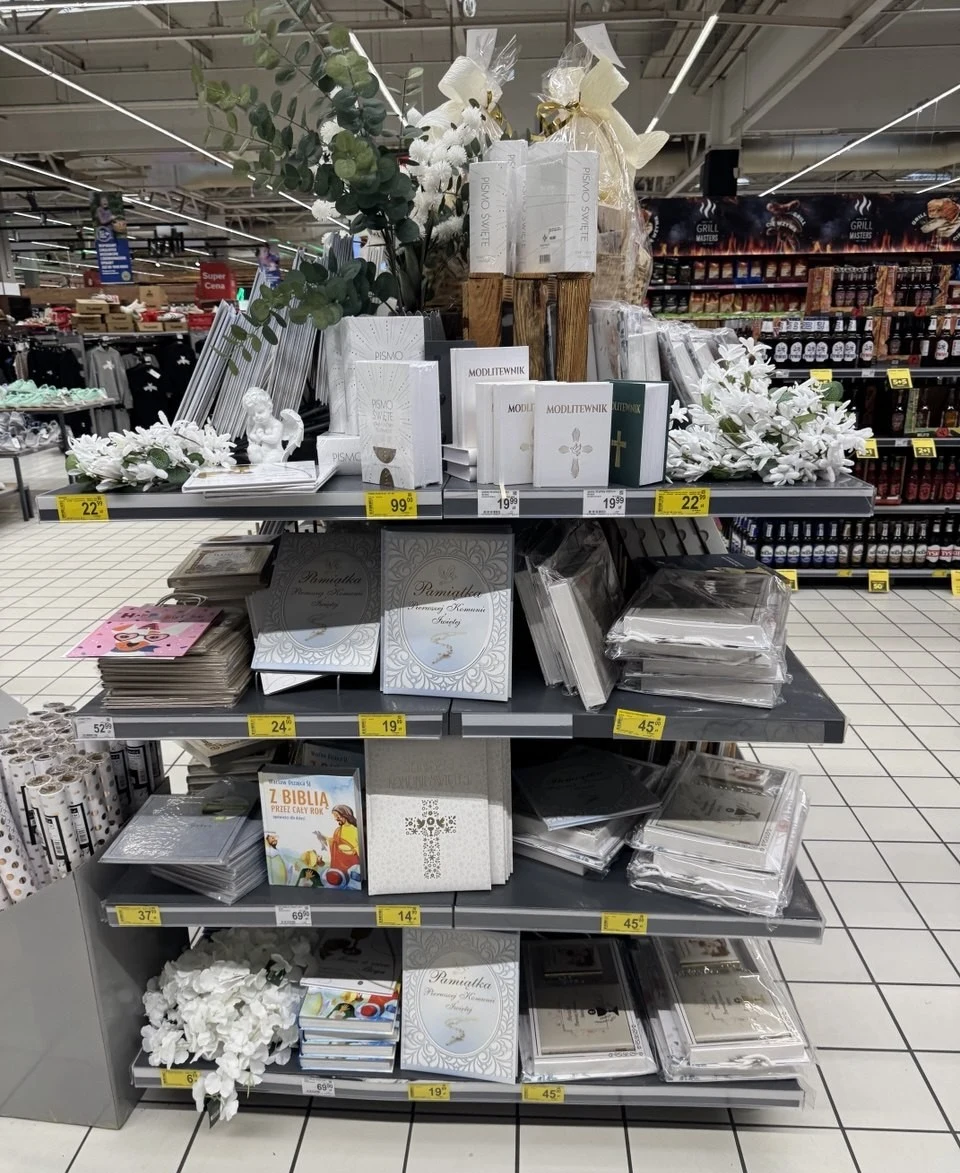 Pamiątki komunijne w Carrefour regał sklepowy z materiałami komunijnymi takimi jak eleganckie albumy, pamiątki pierwszej komunii świętej, białe dekoracje, figurki aniołów oraz książki religijne, w tym biblia dla dzieci; dominują kolory biały i srebrny, podkreślające odświętny charak...
