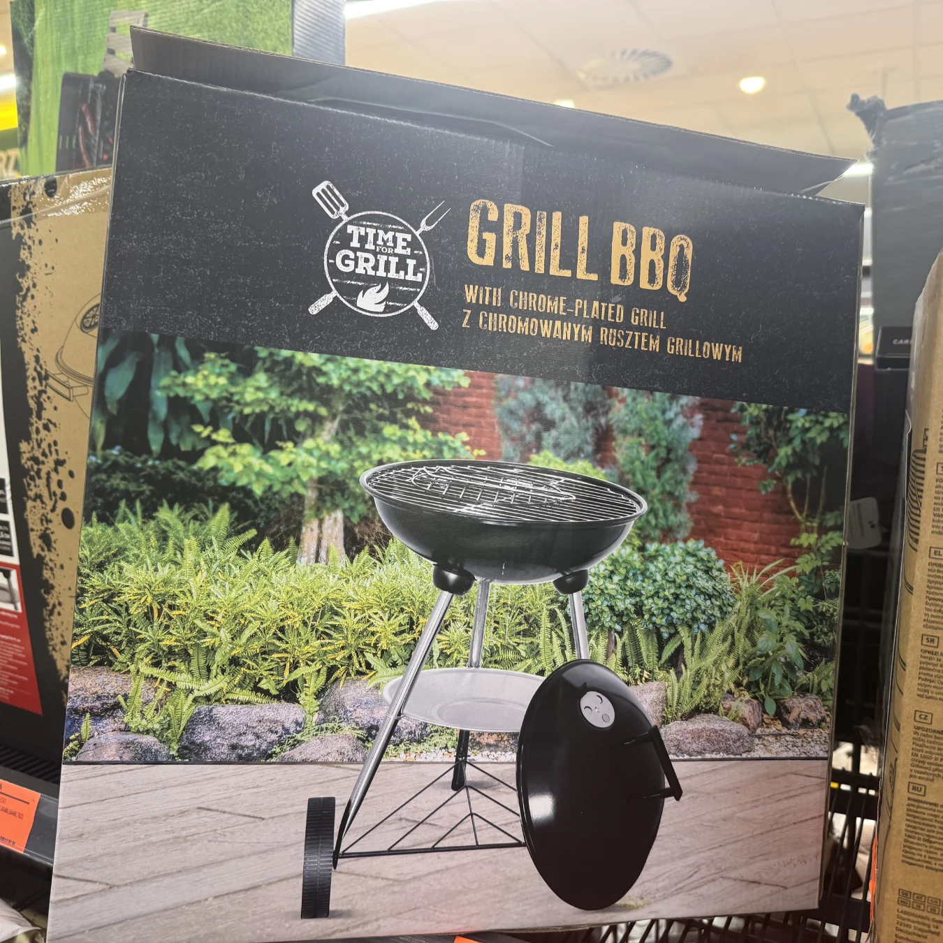 Czarny, okrągły grill na trzech metalowych nogach z chromowanym rusztem, ustawiony na tle ogrodu z zielonymi krzewami i czerwoną ścianą z cegły.