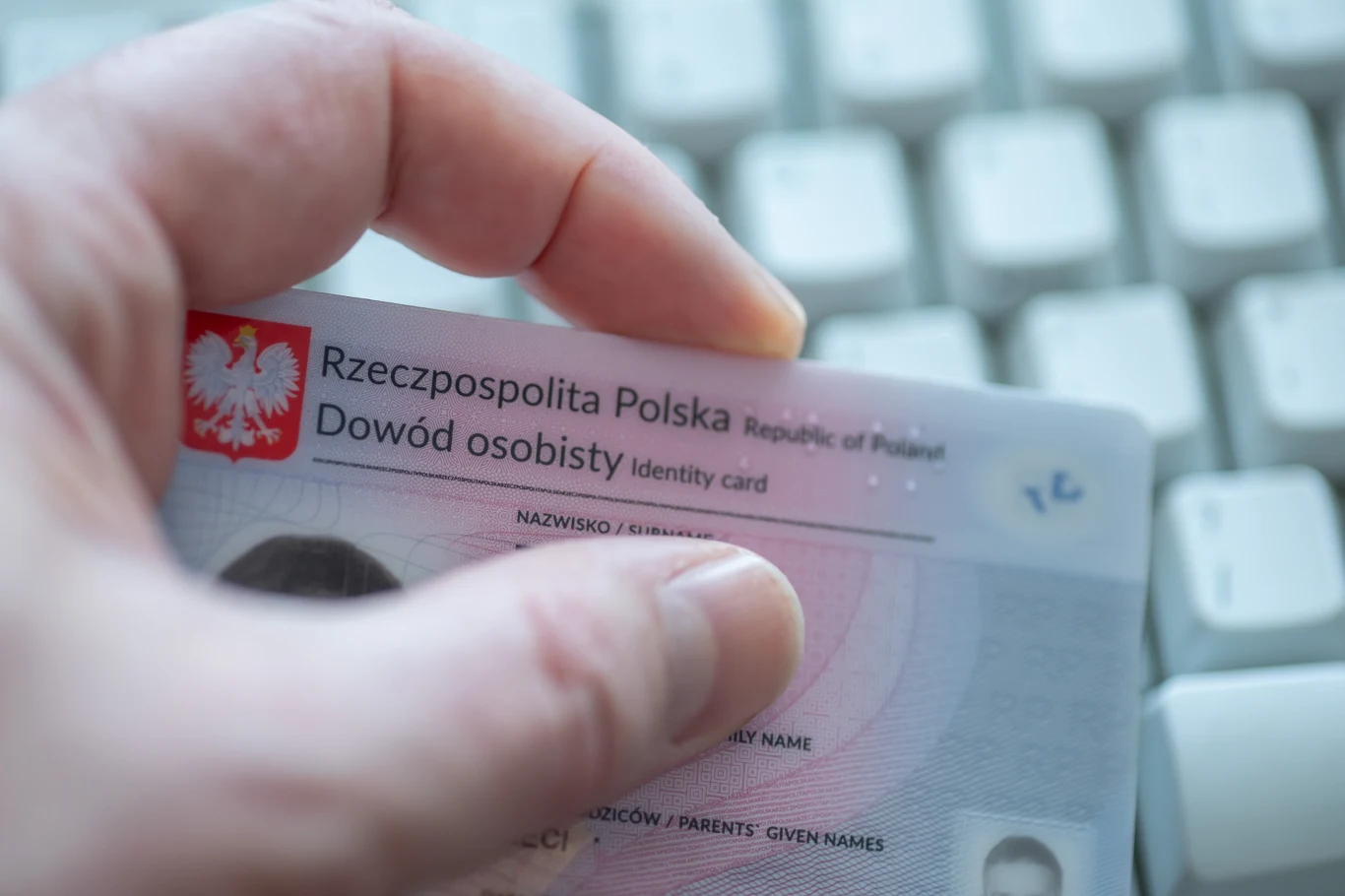 Jak zastrzec dowód osobisty? Jak wyrobić nowy? Ręka trzymająca polski dowód osobisty przed białą klawiaturą komputerową.