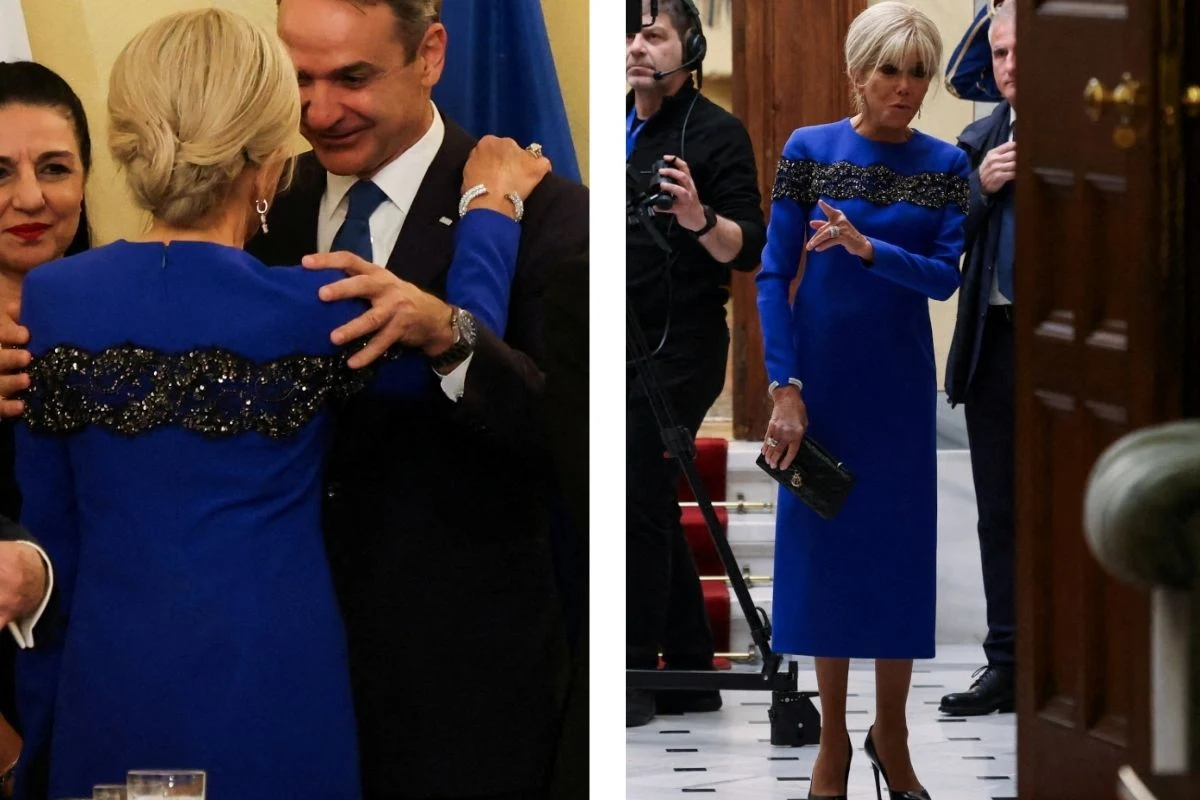 Elegancko ubrana kobieta w niebieskiej sukience z ozdobnym czarnym pasem przechodzi przez korytarz i wita się uprzejmie z mężczyzną w garniturze, w tle obecne są inne osoby oraz fotografowie, cała scena odbywa się w formalnym otoczeniu.