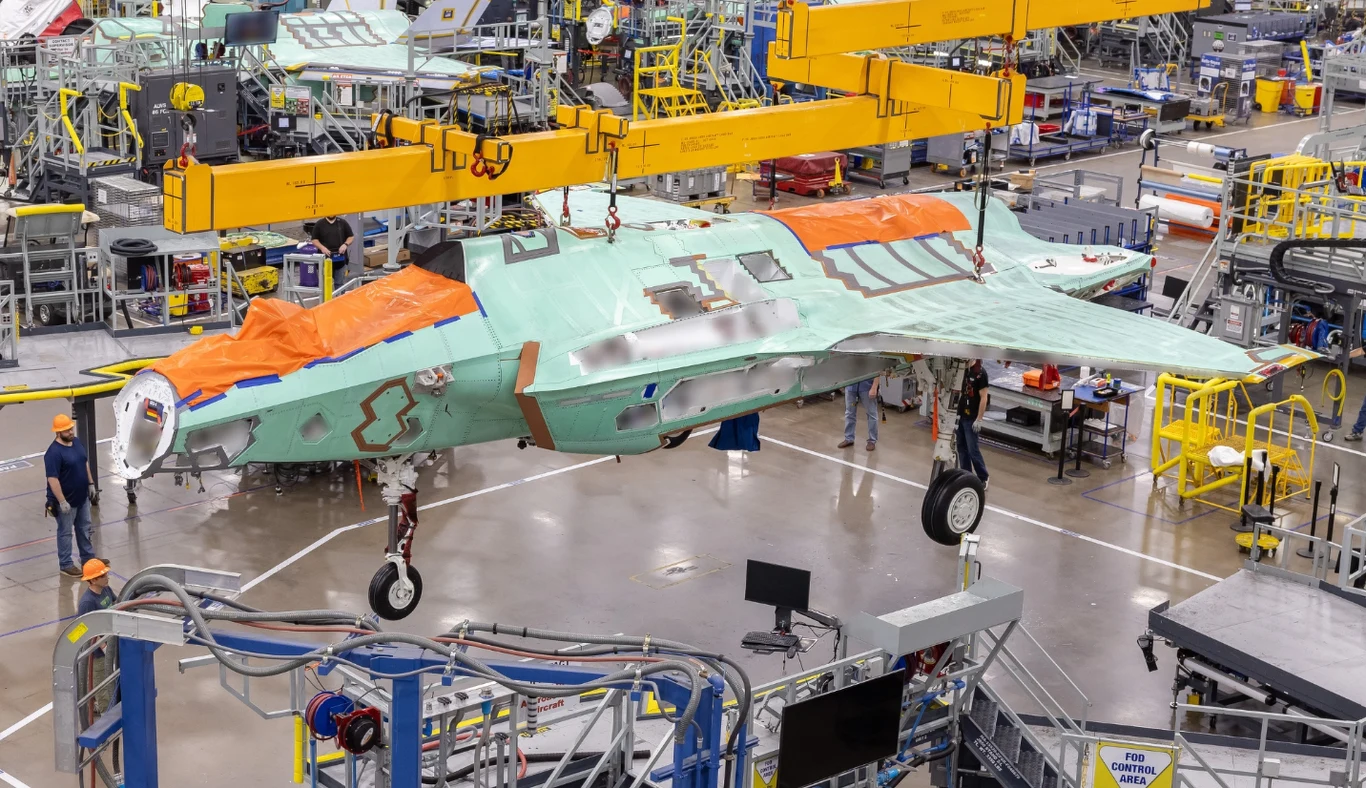 Budowa F-35 dla Niemiec montaż myśliwca na hali produkcyjnej, kadłub samolotu wojskowego zawieszony na mechanicznych dźwigach, intensywnie oświetlone otoczenie z licznymi stanowiskami pracy i konstrukcjami technicznymi, inżynierowie oraz pracownicy w kaskach nadzorujący proces