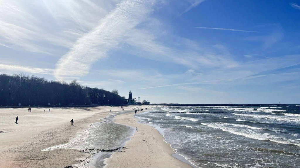 Solo travel w Kołobrzegu to spacery bez mapy szeroka, piaszczysta plaża nad morzem z falami uderzającymi o brzeg, w tle latarnia morska otoczona drzewami oraz pojedyncze osoby spacerujące wzdłuż linii wody pod błękitnym niebem z widocznymi smugami chmur