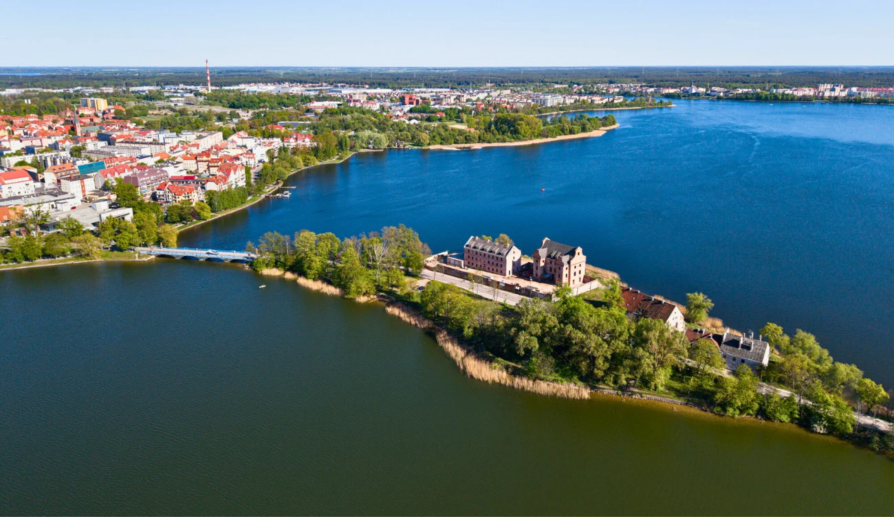 Zamek położony na półwyspie otoczonym wodą, z otaczającymi go zielonymi drzewami i zabudową miejską w tle, w oddali widoczne duże jezioro i panorama miasta.