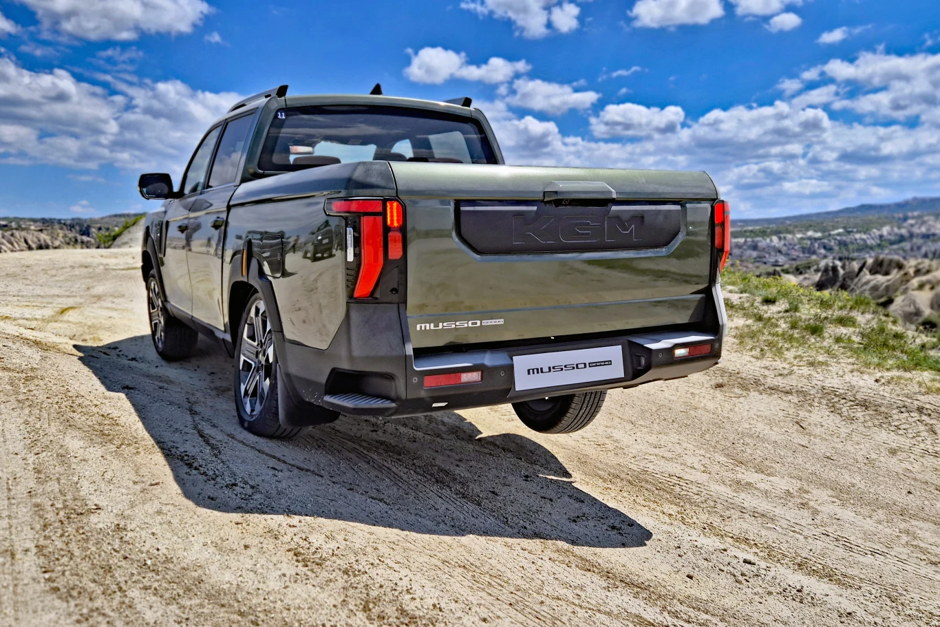 zielony pick-up SsangYong Musso z charakterystycznymi pionowymi tylnymi lampami zaparkowany na piaszczystej drodze w terenie, w tle rozległy krajobraz z pagórkami i błękitnym niebem pełnym chmur