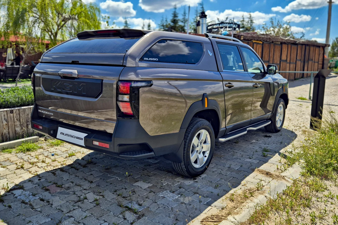 ciemnoszary pickup z zabudowaną paką, nowoczesnym designem tylnych lamp oraz charakterystycznym napisem Musso Grand na klapie; pojazd stoi na brukowanym parkingu w słoneczny dzień