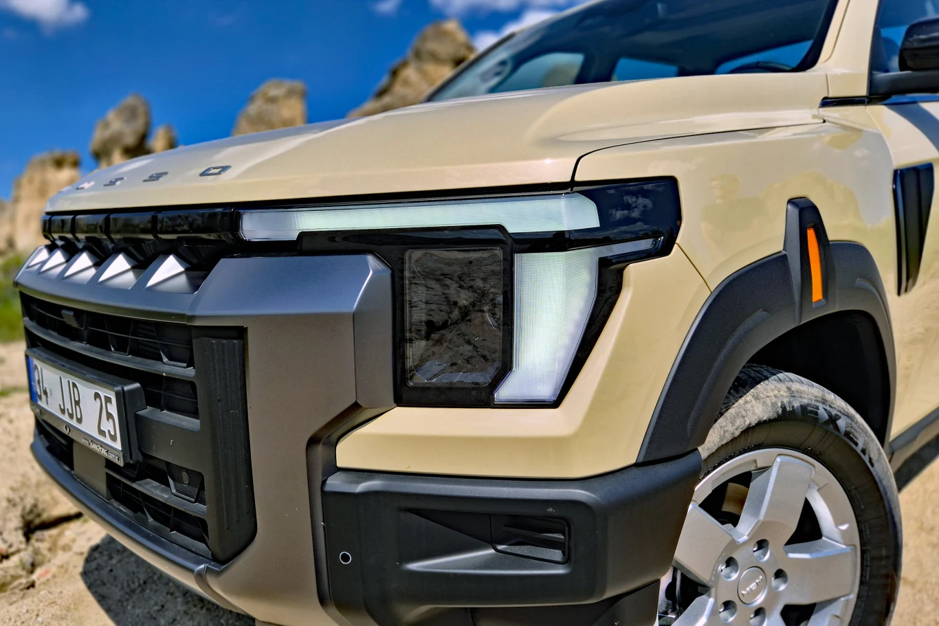 nowoczesny przedni pas beżowego SUV-a z charakterystycznymi, kanciastymi reflektorami LED, masywnym zderzakiem i widocznymi, dużymi felgami na tle jasnego nieba oraz skalistych formacji w oddali