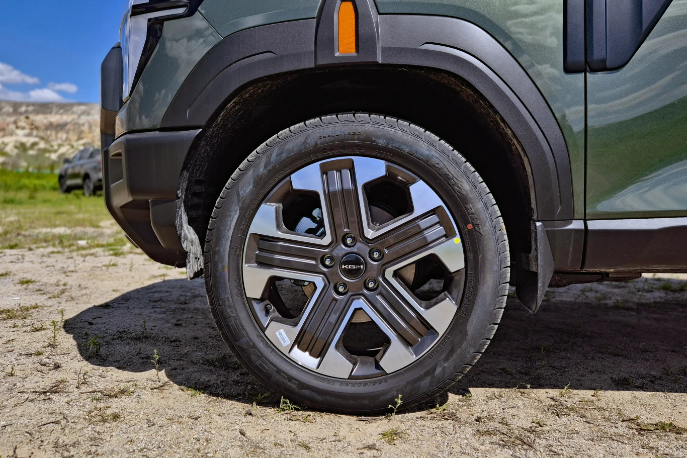 nowoczesna felga aluminiowa z logo Kia na oponie terenowego samochodu, karoseria w kolorze zielonym, wyraźnie widoczne plastikowe osłony nadkoli i elementy reflektora